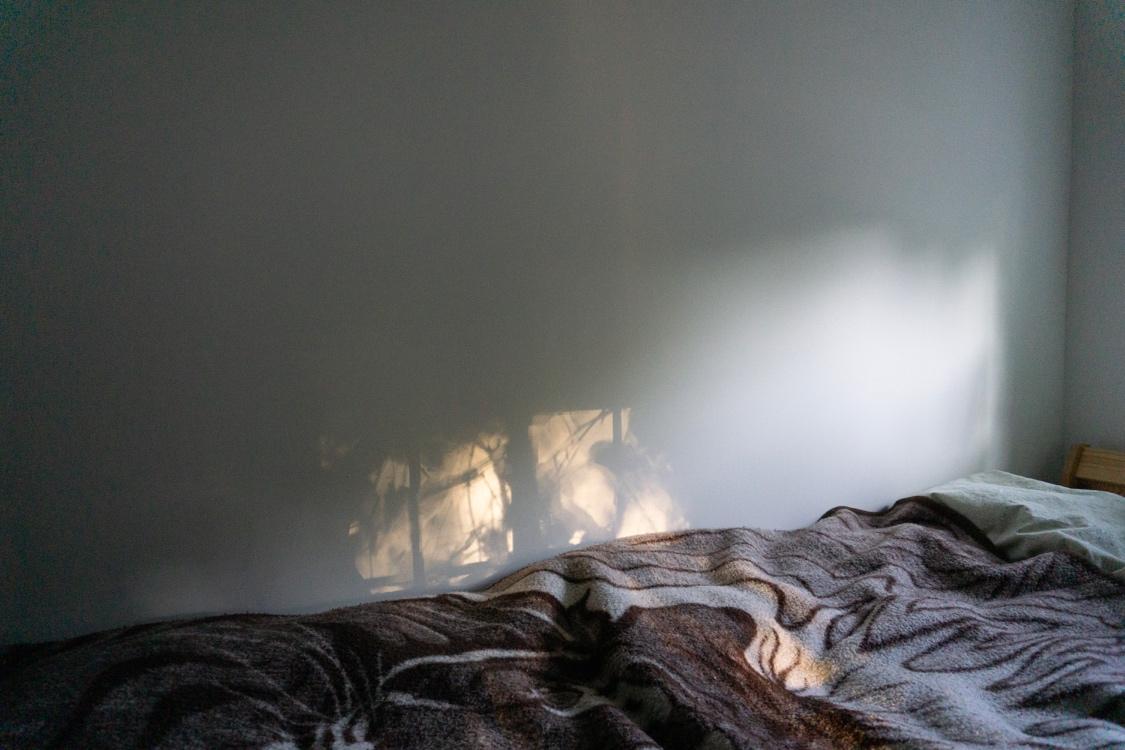 The photograph features a bed in the bottom right corner extending across the bottom of the frame. On the right you can see a pale wooden Ikea frame peeking out, pale green sheets, and a tiger-themed Polish blanket in browns and whites—the thick, winter type. Behind the bed is a bare white wall with glorious light patterns playing on its surface and the surface of the bed, coming from a window on the other side of the room. In this light-window are visible shadows of foliage, the bars of the window, and a bright sunspot. The soft evening creates a warm, soothing ambiance.